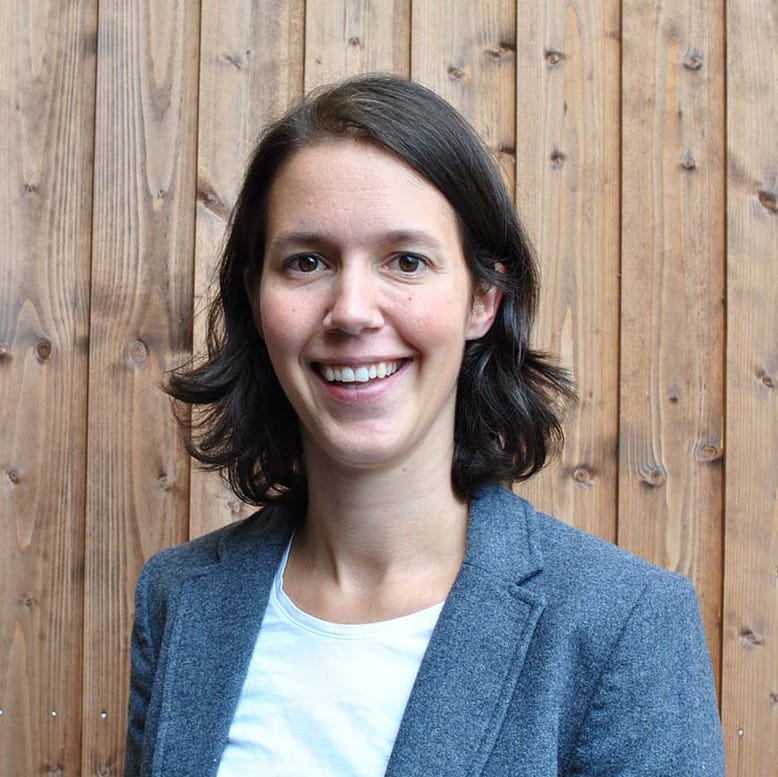 Frau mit dunklem Haar, lächelnd, in grauem Blazer, weißes Hemd, vor einer Holzwand.