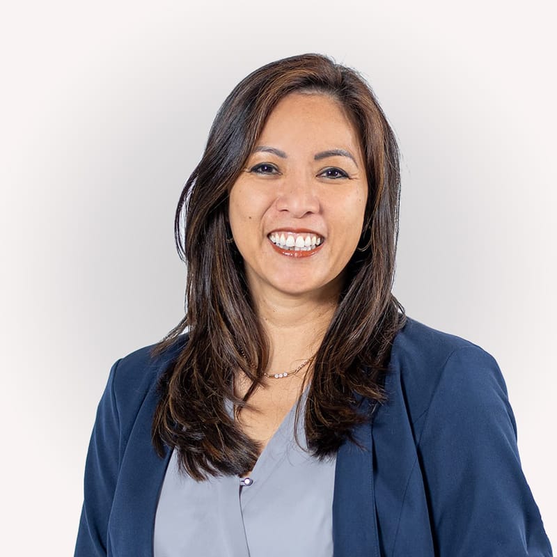 Frau mit langen dunklen Haaren in marineblauem Blazer und grauem Oberteil lächelt in die Kamera.