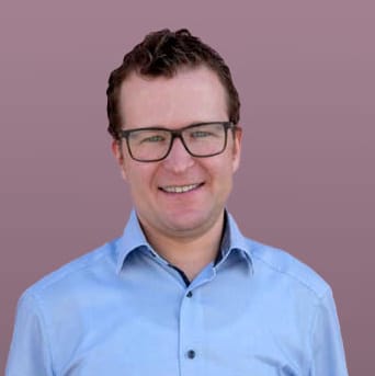 Lächelnde Person mit kurzen braunen Haaren und Brille in blauem Hemd, mauvefarbener Hintergrund.