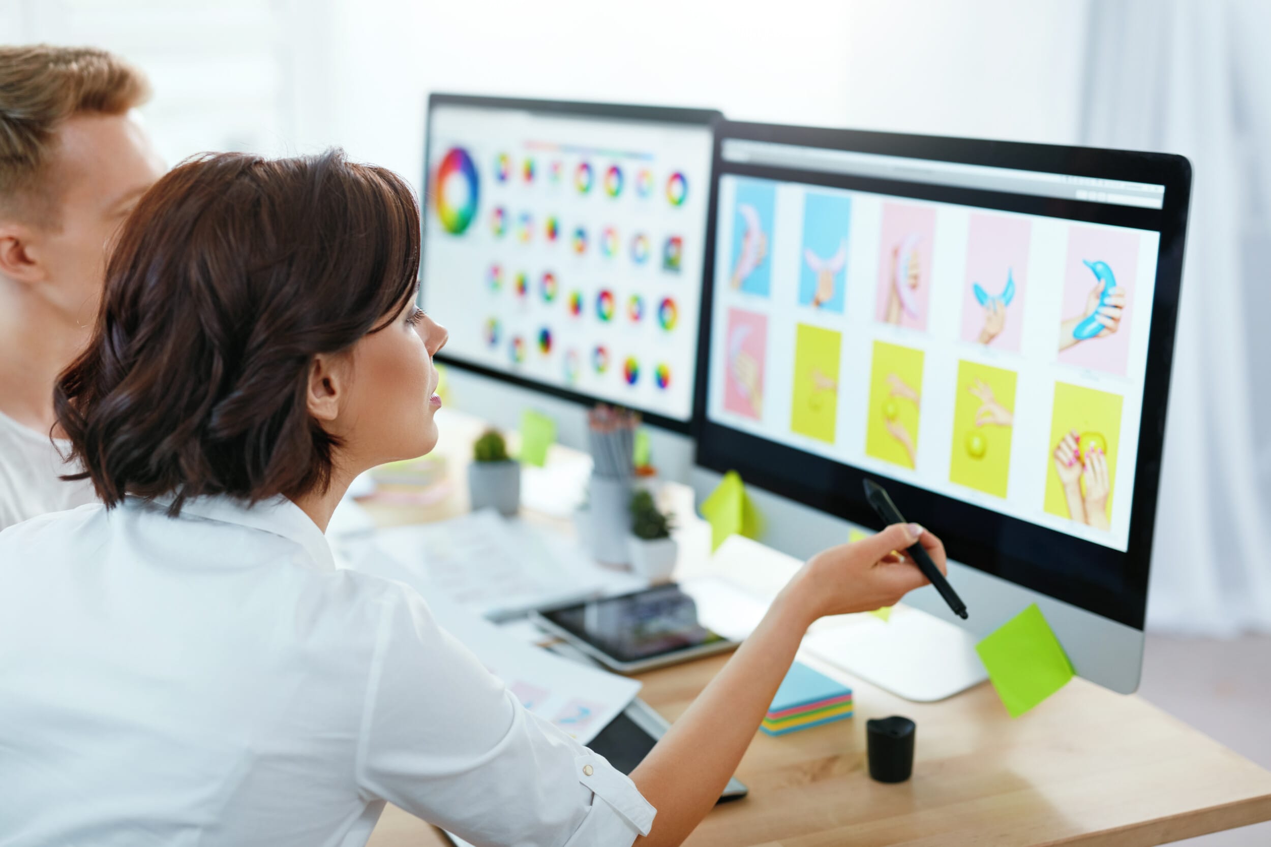 Frau mit Stift an einem Schreibtisch mit zwei Monitoren, auf denen bunte grafische Designs zu sehen sind.