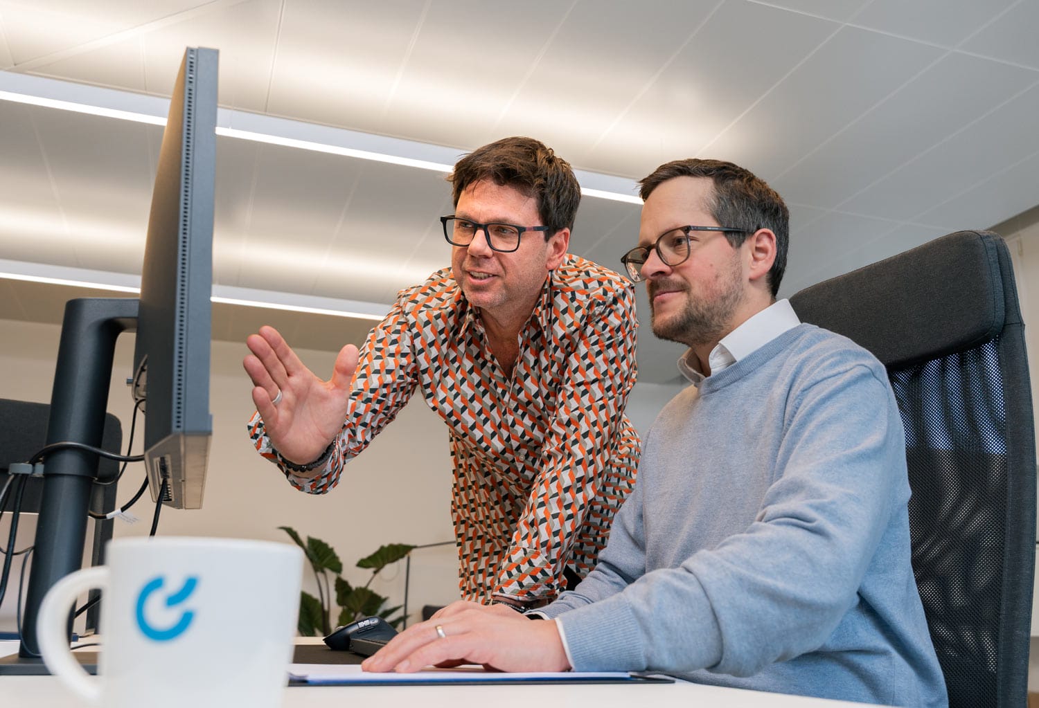 Zwei Männer in einem Büro diskutieren etwas auf einem Computer; Tasse mit blauem Logo davor.