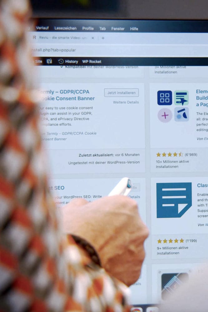 Person hält Smartphone und zeigt auf Computerbildschirm mit WordPress-Plugin-Seite.