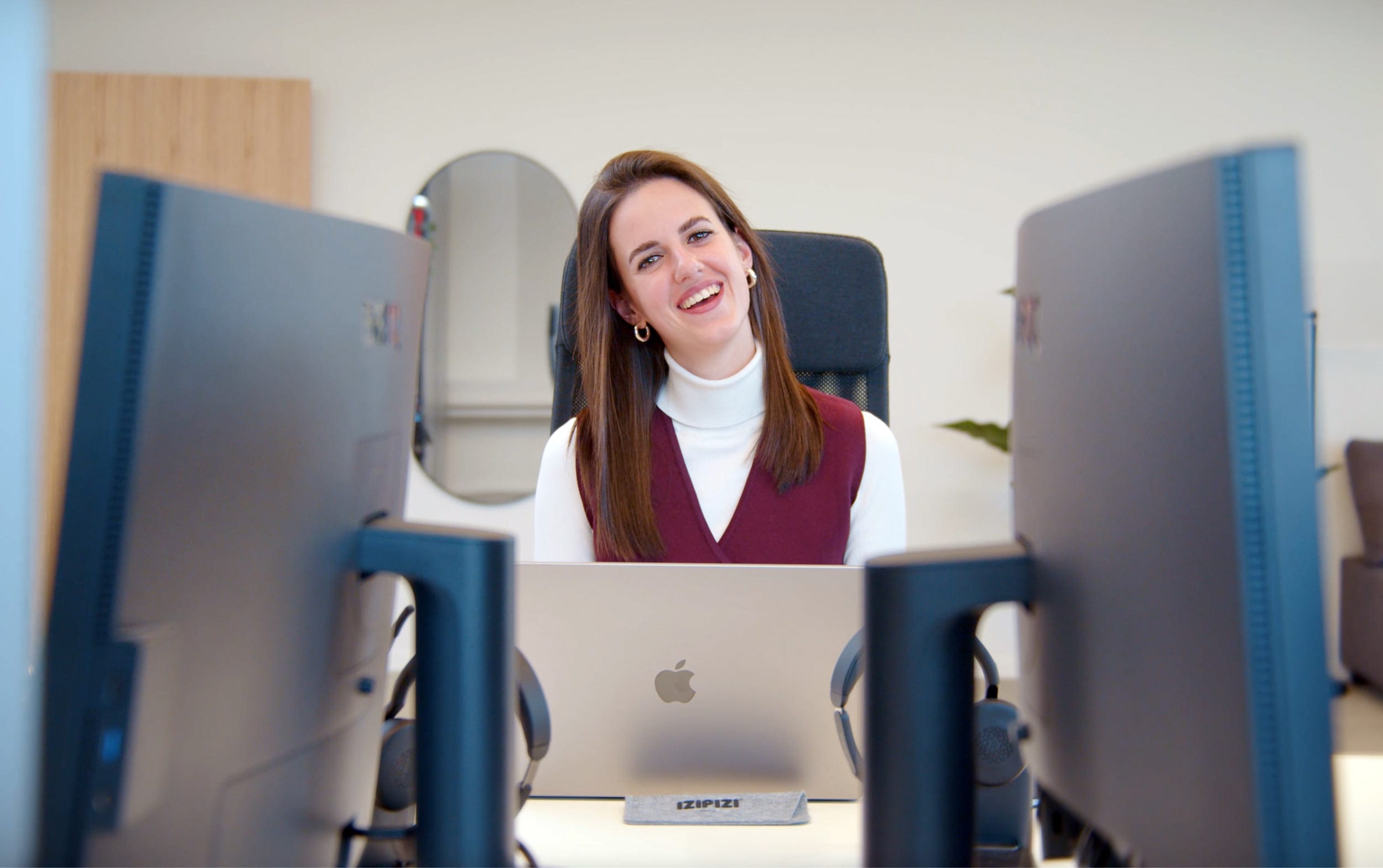 Frau in kastanienbrauner Weste lächelt am Schreibtisch mit Laptop in einem hellen, modernen Büro.