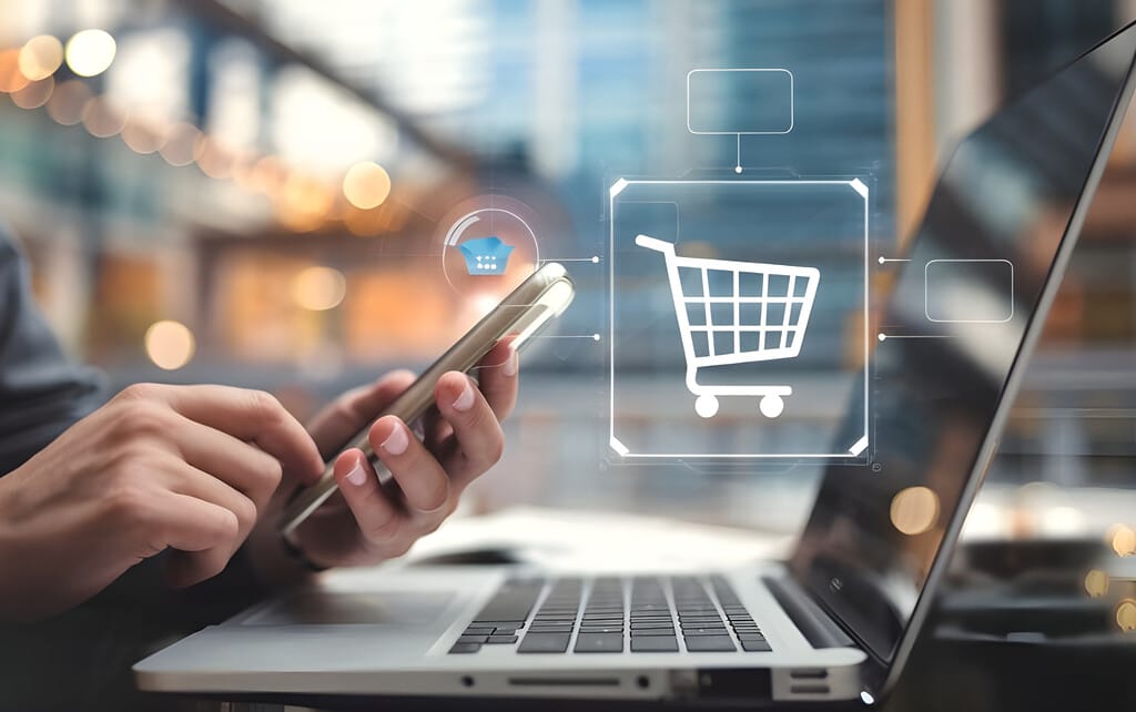 Person mit Smartphone am Laptop, digitale Einkaufskorb-Grafik steht für Online-Shopping.