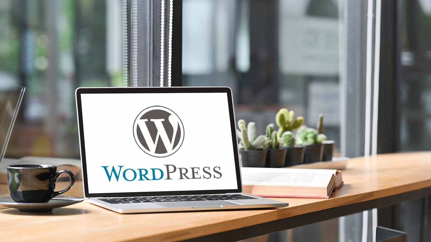 Laptop mit WordPress-Logo auf Schreibtisch neben Tasse, Buch und kleinen Kaktus-Pflanzen.