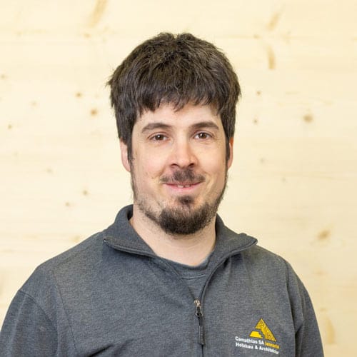 Mann mit kurzen dunklen Haaren und Bart, lächelnd, in grauem Zip-up mit gelbem Logo, Holzhintergrund.