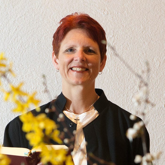 Eine lächelnde Person mit kurzen roten Haaren, schwarzer Jacke und einer Kreuzkette steht vor einer hellen Wand und Blumen.