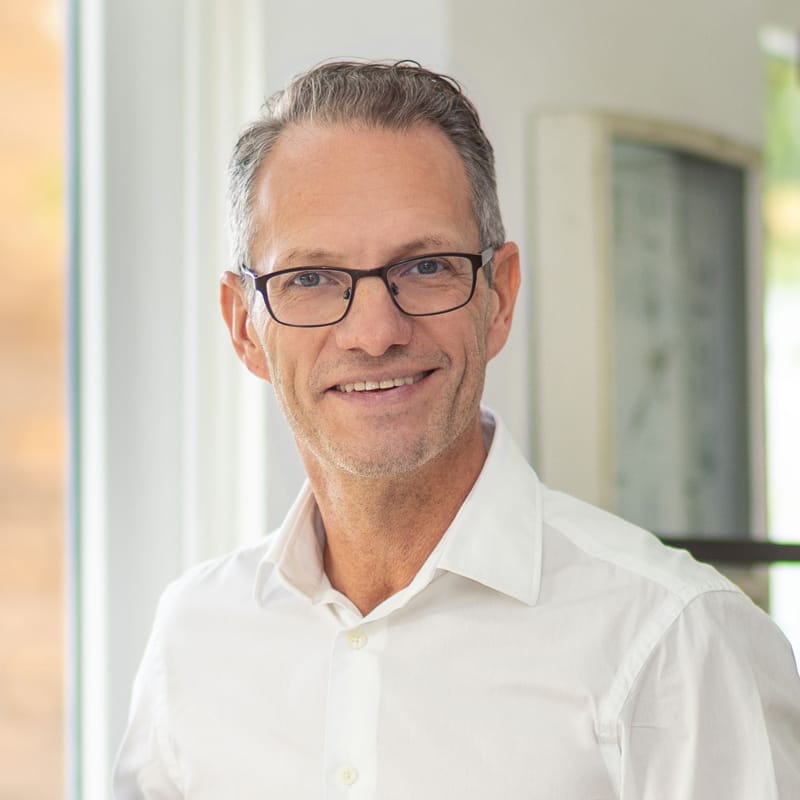 Lächelnder Mann mittleren Alters mit grauen Haaren und Brille in einem weißen Hemd an einem Fenster.